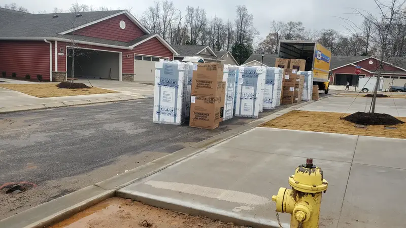 A row of boxed dishwashers staged at the curb in a new subdivision, with a rental box truck at the far end