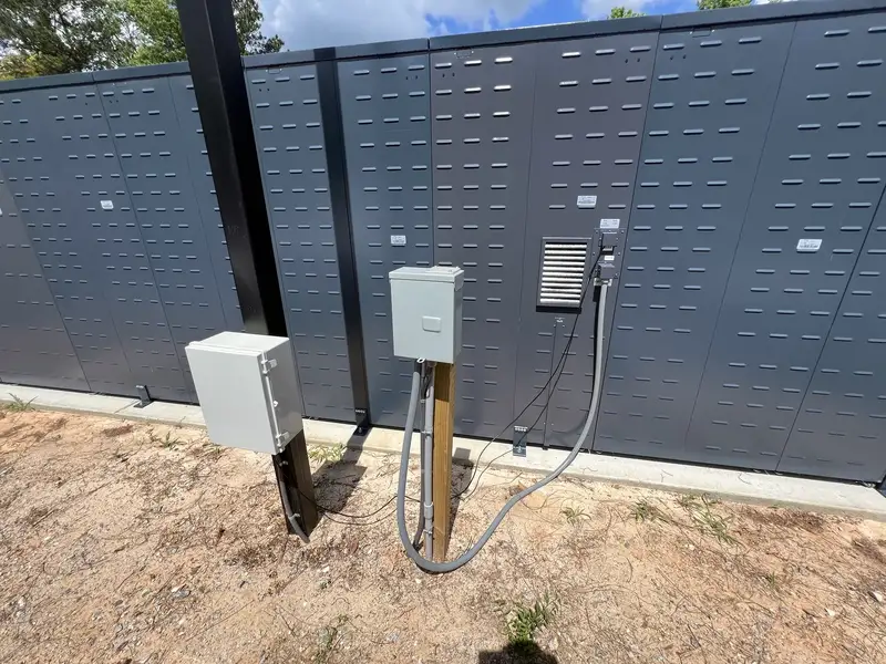 Back of an outdoor locker row showing junction boxes, flexible conduit and posts on a new concrete pad under a blue sky