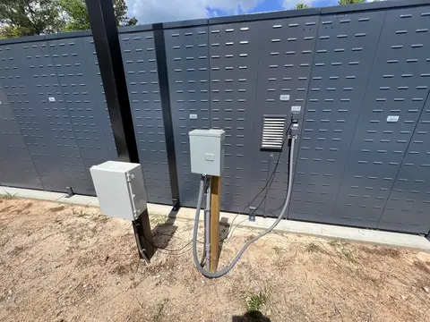 Back of an outdoor locker row showing junction boxes, flexible conduit and posts on a new concrete pad under a blue sky