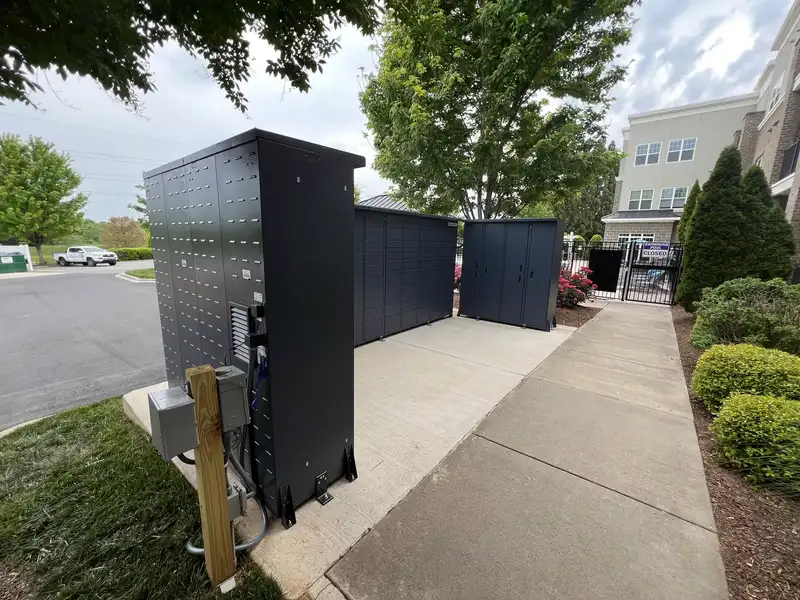 Three charcoal locker units on a new concrete pad with a wooden power and data post and conduit in front, at an apartment complex