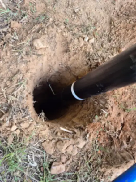 Top-down view of a black pole with a white reflective band being set in a freshly dug round hole in red soil
