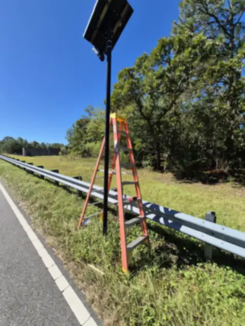 Black pole with a solar panel and a camera beside a roadside guardrail, with an orange stepladder leaning on it