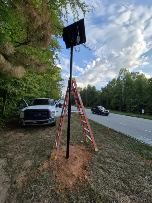 Solar highway camera pole freshly set beside a road, with a ladder against it and a work truck parked behind