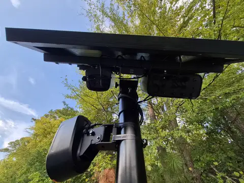 Looking up at a black pole with a solar panel on top and two cameras mounted below it, against a blue sky